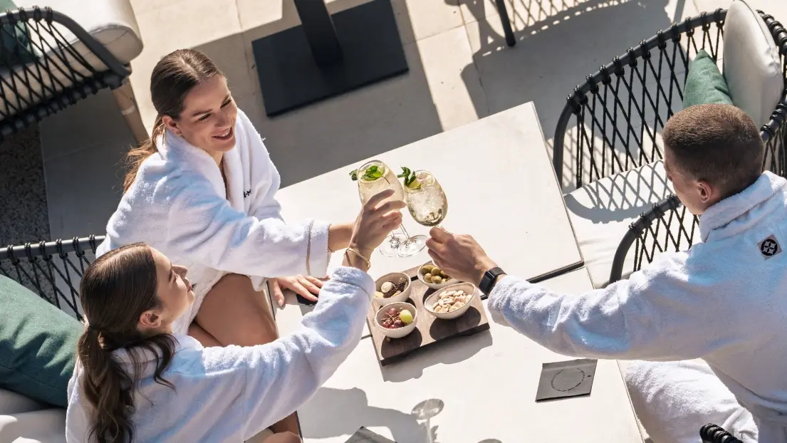 Three friends in bathrobes sip drinks and snacks by the pool.