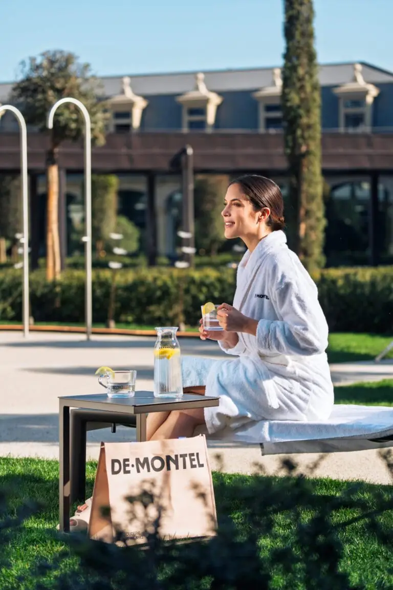 Woman relaxing in a bathrobe by the pool, sipping a drink. De-Montel bag visible.