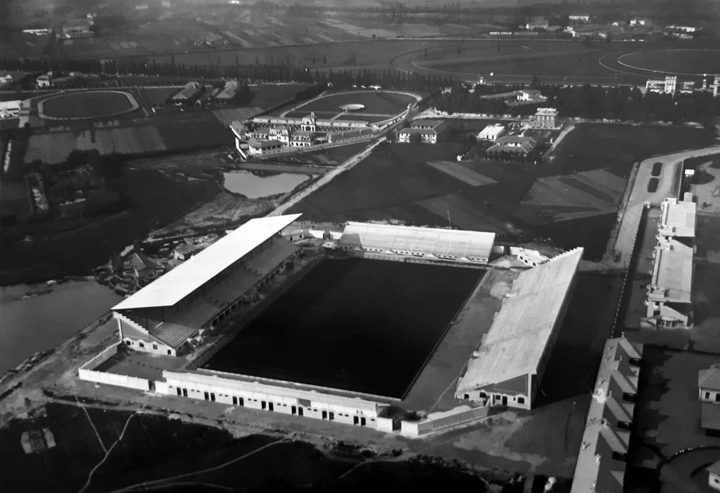 Vista aerea di uno stadio in costruzione, circondato da edifici e campi circostanti.